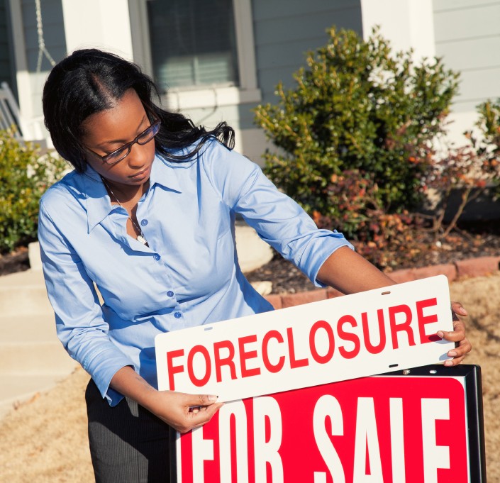 A woman putting a FORECLOSURE sign on a red sign that says FOR SALE referring to a house, positing the question, How does foreclosure work in Georgia and Athens?